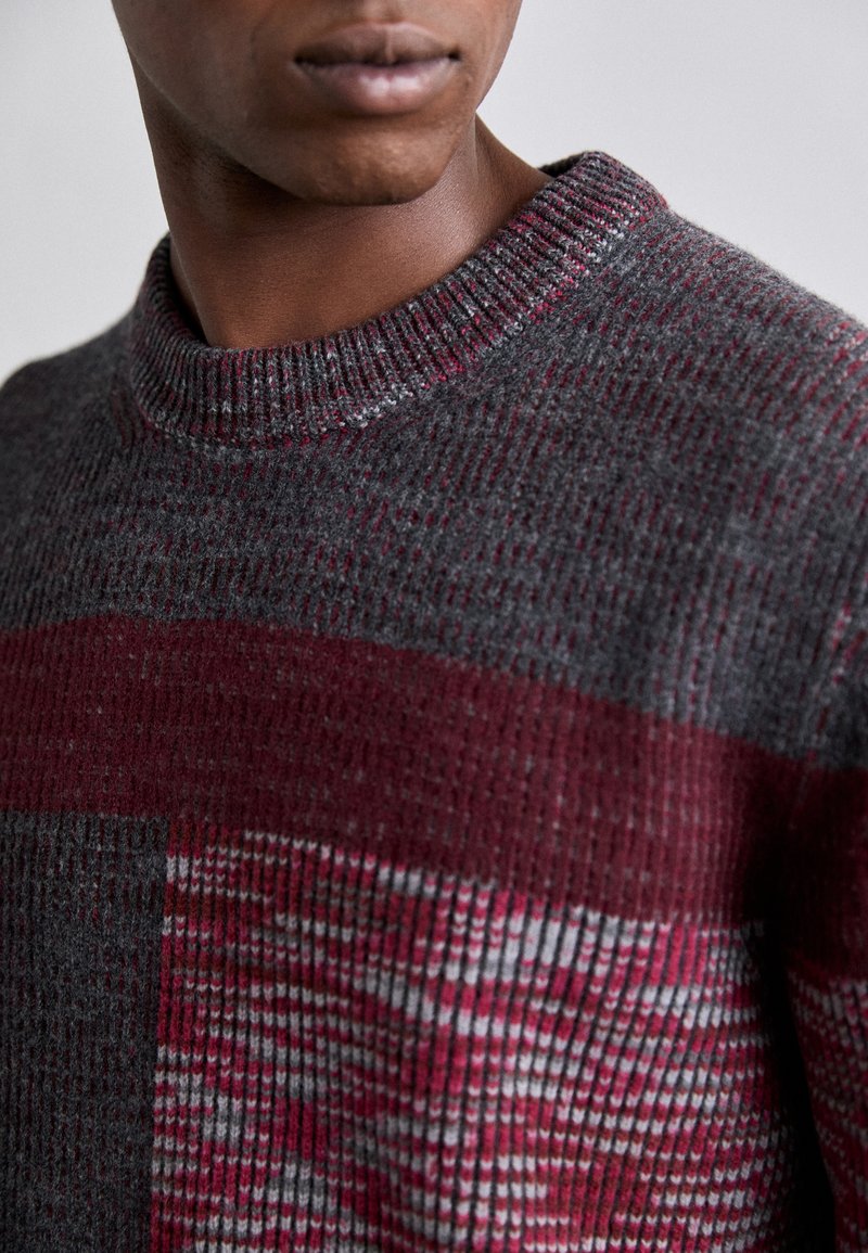 Knitted sweater in dark gray with maroon and lighter gray horizontal stripes. Ribbed neckline and textured fabric with a pattern.