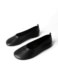 Fekete bőr slip-on lapos cipő páros, lekerekített orral és kis sarkú fülekkel, fehér háttéren.