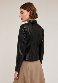Giacca in pelle nera dal taglio slim, con colletto rialzato, cuciture sulle spalle e hardware minimale. Abbinata a una gonna beige.