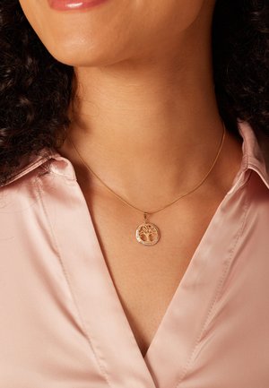 Collier en or avec un pendentif circulaire au design d'arbre, encadré d'une bordure texturée, porté sur un chemisier en satin rose clair.