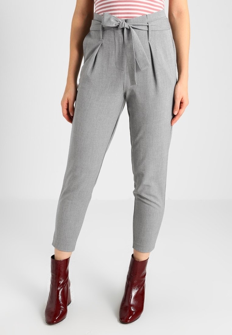 Femme portant un pantalon gris taille haute et fuselé avec une ceinture en tissu nouée en noeud, associé à des bottines bordeaux et un top à rayures roses.