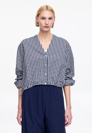 Kobieta w granatowo-białej, kraciastej bluzce na guziki z podwiniętymi rękawami i granatowych spodniach, stojąca na białym tle.