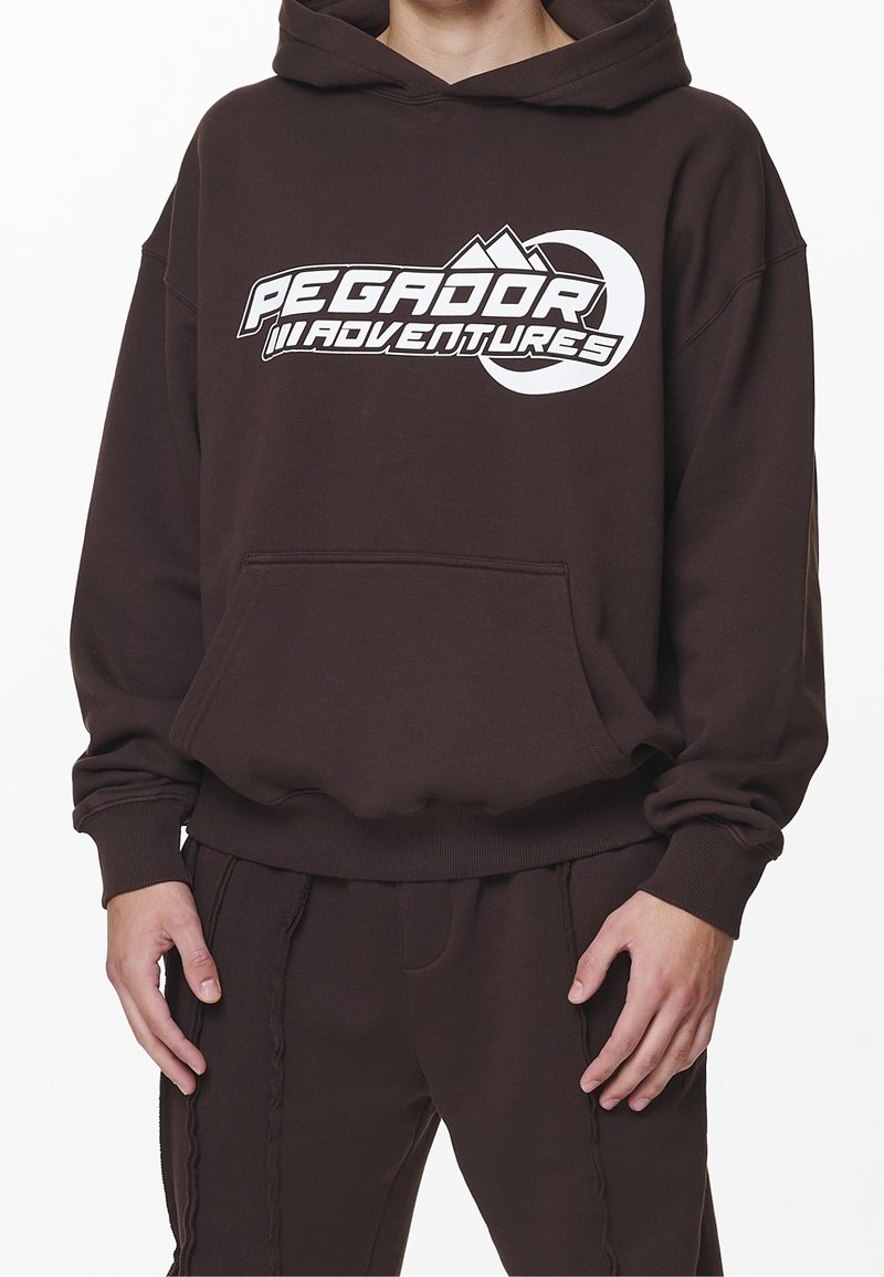 Bruine hoodie met een groot wit "PEGADOR ADVENTURES" logo, een kangaroo zak en geribbelde manchetten en zoom. Gecombineerd met bijpassende broek.
