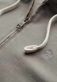 Gray zip-up hoodie featuring a textured fabric, gold zipper, and white drawstring with a logo embroidered in white on the chest.