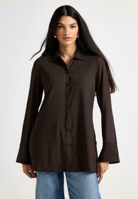 Camicia marrone a maniche lunghe con bottoni e colletto, vestibilità slim e polsini svasati. Presenta una texture liscia e una leggera lucentezza. Indossata con jeans azzurri.