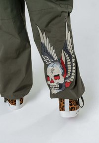 Pantaloni cargo verde oliva con un design di teschio e ali ricamato. Sneakers marroni con stampa leopardata e suole bianche.