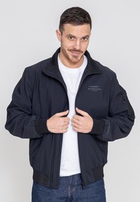 Schwarze Bomberjacke aus glattem Stoff, mit hohem Kragen, Reißverschluss-Taschen, gerippten Bündchen und einem Logo auf der oberen linken Seite.