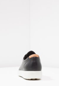 Schwarzer Lederschuh mit strukturierter Oberfläche, weißer Gummisohle und braunem Akzent an der Ferse. Einfaches Design, runde Form und flache Profile.