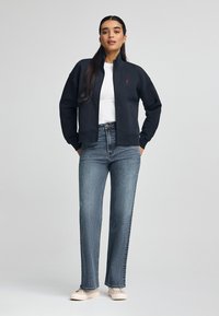 Giacca nera con zip sopra una maglietta bianca, abbinata a jeans blu a vita alta e sneakers beige. Presenta un piccolo logo rosso sulla giacca.