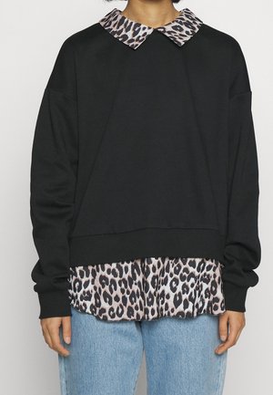 Personne portant un pull noir à manches longues sur une chemise à col imprimé léopard, associé à un jean en denim bleu clair, se tenant contre un fond uni.