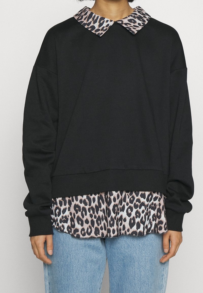 Person som har på sig en svart långärmad tröja över en leopardmönstrad skjorta med krage, tillsammans med ljusblå jeans, står framför en neutral bakgrund.