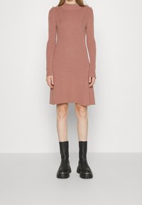 Robe en maille côtelée rose poudré avec un col haut, des manches longues et des accents dorés aux épaules ; portée avec des bottines noires épaisses.