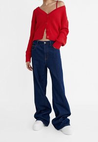 Roter gerippter Cardigan mit fünf Knöpfen und weiten Ärmeln, kombiniert mit hoch taillierten, locker sitzenden dunkelblauen Jeans. Weiße Plateauschuhe.