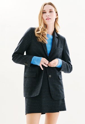 Geruite blazer in marineblauw en donkerblauw, met twee voorvakken en een enkele knoopsluiting, gecombineerd met een lichtblauwe trui en rok.