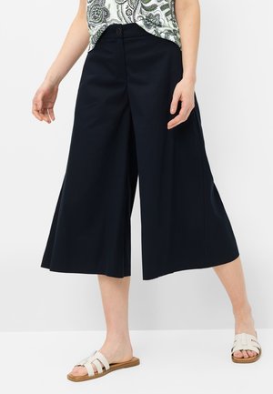 Femme portant un pantalon noir large et court, un haut blanc à motifs, et des sandales blanches à enfiler, debout sur un fond blanc.
