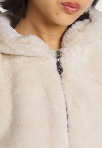 Kremowa kurtka z futerka ekologicznego z kapturem i srebrnym zamkiem błyskawicznym. Charakteryzuje się miękką teksturą i dopasowanym krojem, podkreślającym puszysty materiał.