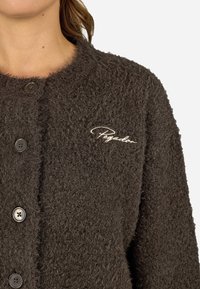 Hnědý, chlupatý kardigan s knoflíky a vyšitým logem "Pegador" na hrudi. Měkká textura a volný střih.