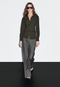 Dark green button-up shirt with long sleeves, paired with gray tailored pants. Model wearing black sunglasses and pointed heels, standing on a black backdrop.