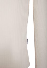 Vaalea beige ribattu paita pitkillä hihoilla, jossa on suora leikkaus ja pieni etiketti vasemmassa saumurissa. Valmistettu pehmeästä kankaasta.