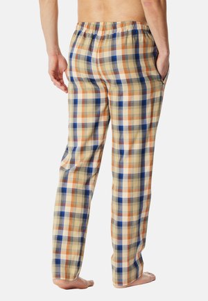 Person stående barfodet iført beige, orange og blåternede pyjamasbukser med elastisk linning og hænder i lommerne, vist bagfra.