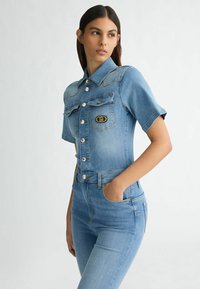 Denim-Overall in Hellblau, mit kurzen Ärmeln, Knopfleiste, Brusttaschen und einem Logopatch. Figurbetonter Schnitt mit glatter Textur.