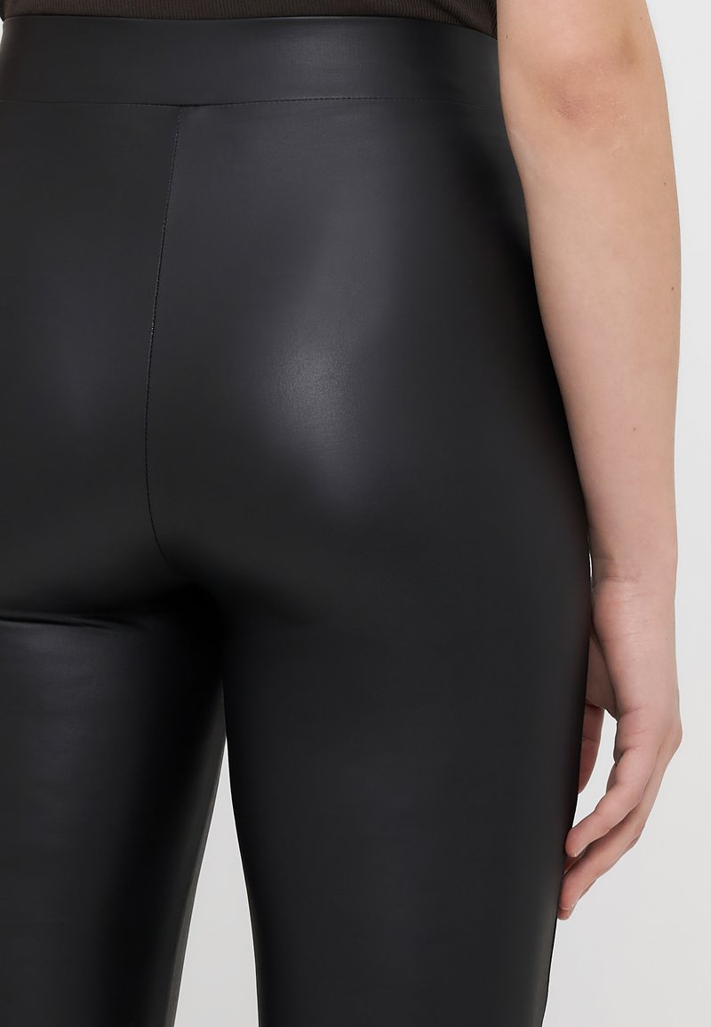 Schwarze Leggings aus Kunstleder mit glatter Textur, hohem Bund und figurbetontem Design, die die Rückansicht und einen Teil der rechten Hand zeigen.