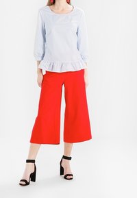 Blouse à rayures bleu clair avec un ourlet peplum, associée à un pantalon large rouge vif. L'ensemble est complété par des sandales à talons blocs noires avec des lanières.