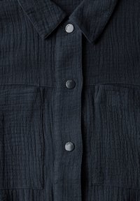 Chemise texturée bleu marine avec une fermeture à boutons sur le devant, un col classique et deux poches poitrine. Présente des boutons ronds noirs avec le nom de la marque "Cecil".