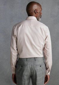Helles rosafarbenes Langarmshirt mit glatter Textur, kombiniert mit grauen karierten Hosen. Auffällige Knopfdetails an den Ärmeln und am Rücken.