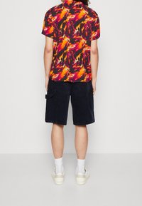 Lyhythihainen paita, jossa on eloisa abstrakti kuvio oranssina, punaisena ja mustana; yhdistettynä tummiin samettishortseihin ja valkoisiin lenkkareihin.