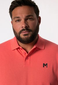 Korálová polo košile s límečkovým designem. Obsahuje tři knoflíky a malé černé logo "M" na levé části hrudníku. Měkká, strukturovaná tkanina.