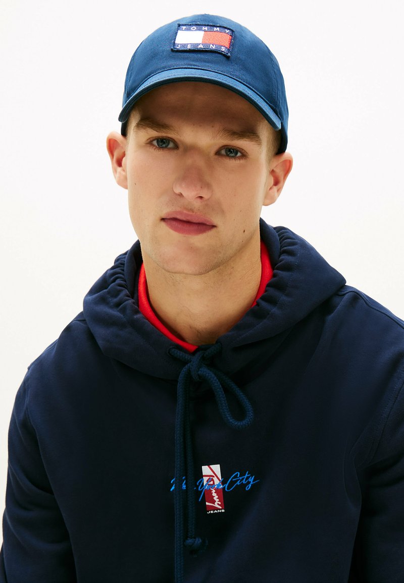 Młody mężczyzna w granatowej bluzie z kapturem z czerwonym logo i dopasowaną czapką Tommy Jeans, patrzący prosto w obiektyw.