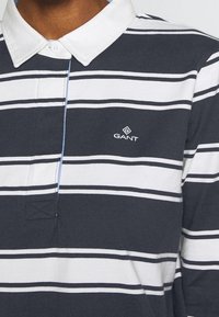 Dunkelblaues und weiß gestreiftes Poloshirt mit einem weißen Kragen, das ein kleines GANT-Logo auf der Brust trägt. Aus weichem Baumwollmaterial mit legerem Schnitt.