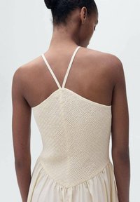 Das cremefarbene Kleid hat ein gerafftes Oberteil mit dünnen Trägern und einen schwingenden Rock, der die Textur und sanfte Drapierung betont.