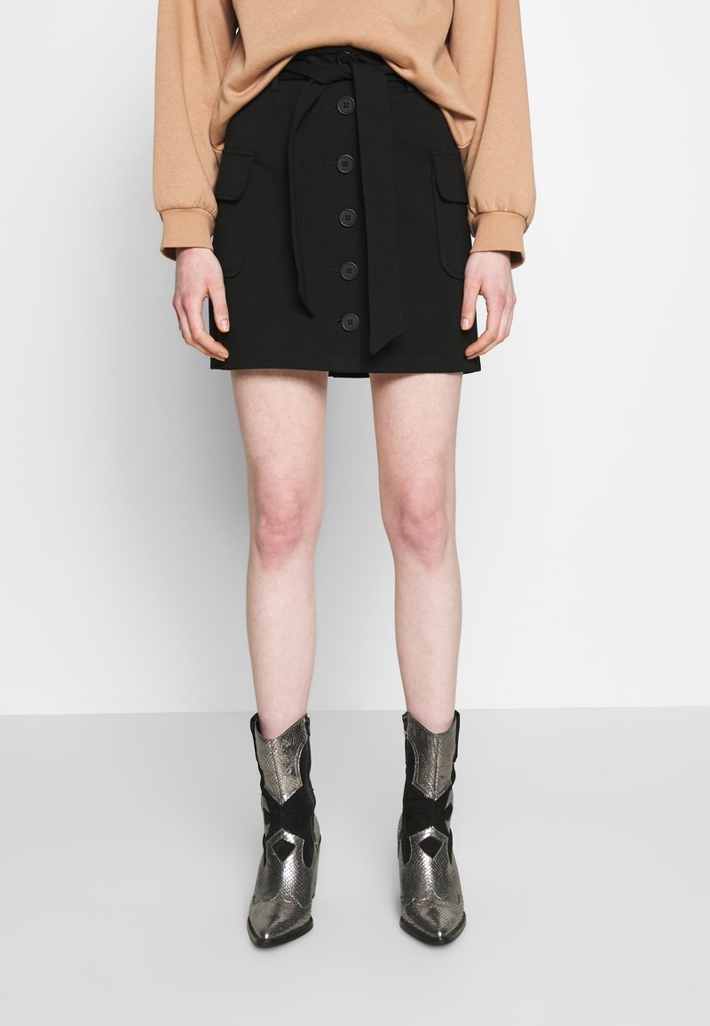Femme portant une jupe noire boutonnée devant avec poches et ceinture à nouer, un haut à manches longues beige, et des bottines métalliques argentées et noires.