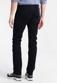Dunkelblaue Jeans aus Denim mit schlanker Passform, fünf Taschen-Design und subtiler Verblassung. Getragen mit schwarzen und olivgrünen Sneakers.