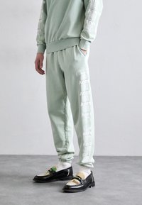 Lys grøn sweatshirt og sweatpants med hvid "Aries" branding langs siden. Sorte loafers med farverige tekstil accenter og stropdetalje.