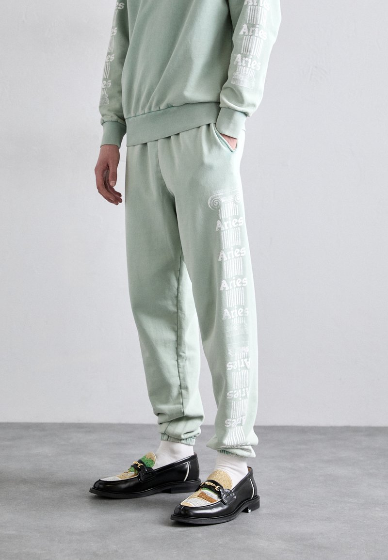 Lys grøn sweatshirt og sweatpants med hvid "Aries" branding langs siden. Sorte loafers med farverige tekstil accenter og stropdetalje.