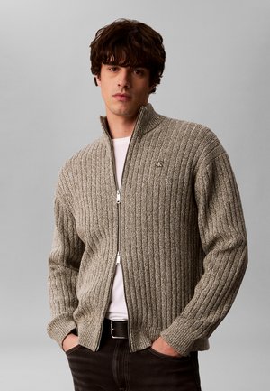 Junger Mann trägt einen beigen, gerippten Reißverschluss-Pullover über einem weißen Hemd mit den Händen in dunklen Hosentaschen vor schlichtem Hintergrund.