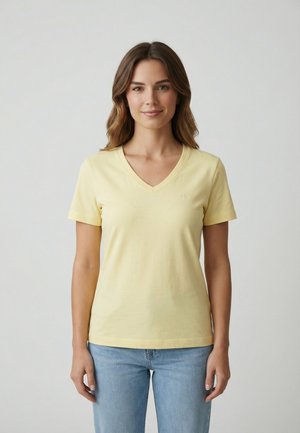 Mujer con cabello castaño ondulado, vestida con una camiseta de cuello en V de color amarillo pálido y jeans azul claro, posando frente a un fondo blanco liso.