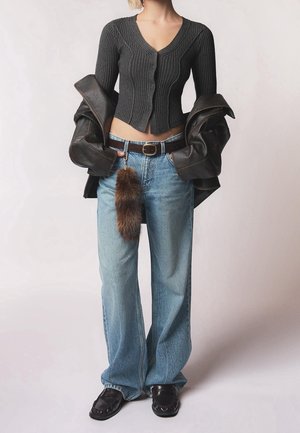 Frau trägt ein kurzes dunkelgraues Cardigan, weite hellblaue Jeans mit braunem Gürtel und pelzigem Schlüsselanhänger, hält eine schwarze Lederjacke.