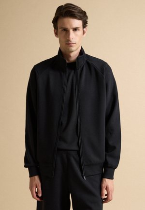 Giovane uomo con capelli corti e castani indossa una giacca nera con zip sopra una maglietta nera, in piedi davanti a uno sfondo beige.