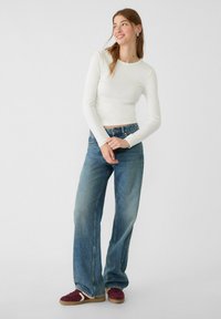 Weißes, langärmliges, figurbetontes Oberteil, kombiniert mit weit geschnittenen blauen Jeans. Das Model trägt bordeauxrote Sneakers mit hellfarbiger Sohle. Einfaches, modernes Design.