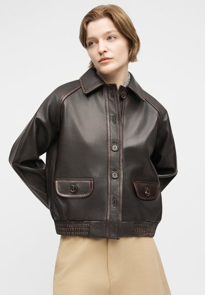 Νεαρό άτομο με κοντά μαλλιά φοράει σκούρο καφέ δερμάτινο σακάκι με κουμπιά και τσέπες με καλύμματα, στέκεται μπροστά σε απλό φωτεινό φόντο.