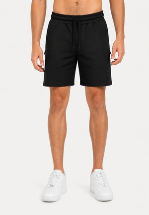 Homme portant un short noir à cordon et des baskets blanches, debout devant un fond clair uni.