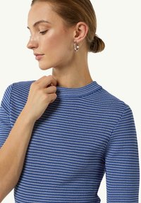 Blue and white striped long-sleeve top with a high collar, featuring a textured fabric and a fitted silhouette. Silver hoop earrings are worn.