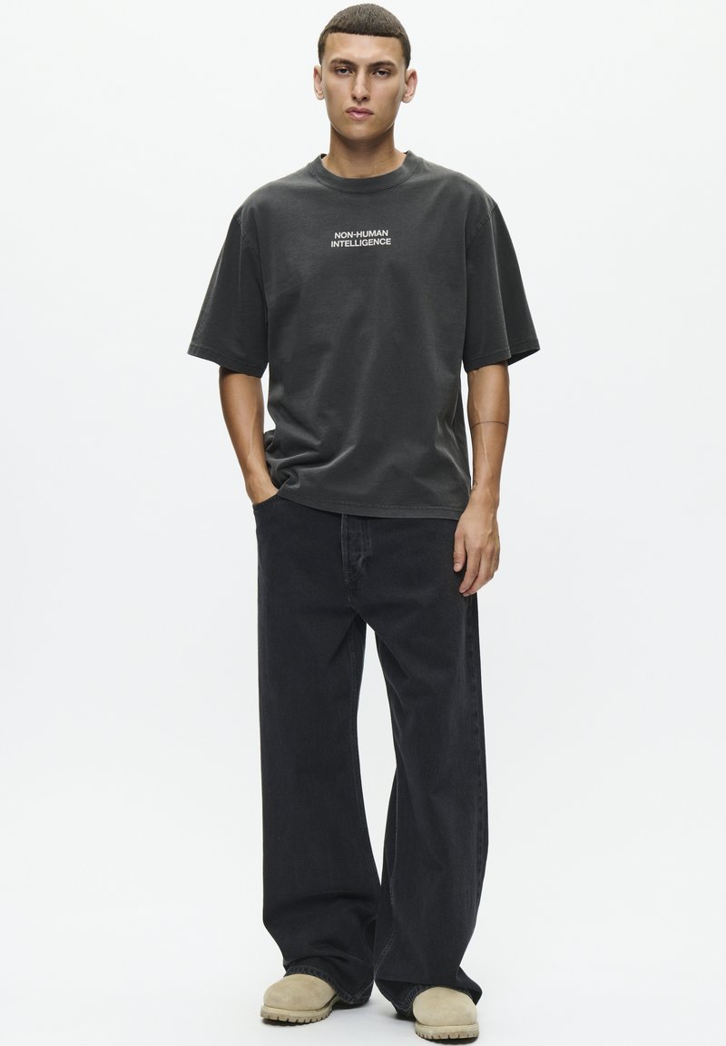 T-shirt en coton gris avec le texte "INTELLIGENCE NON HUMAINE", associé à un jean noir à jambe large et des sabots crème. Design simple et coupe décontractée.