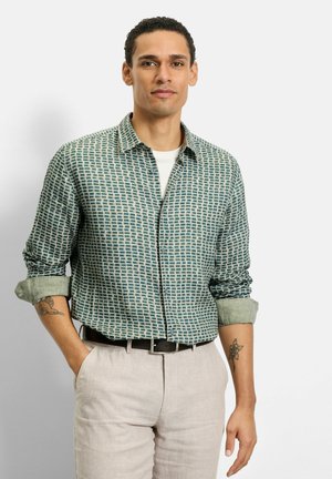 Mann trägt gemustertes Langarmhemd mit hochgekrempelten Ärmeln, hellfarbene Hose, schwarzen Gürtel und sichtbare Tätowierungen auf beiden Unterarmen, steht vor weißer Wand.