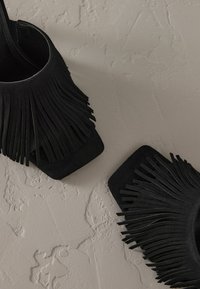 Sandales à talons en daim noir avec des détails en franges, un bout carré et un design de bride minimaliste à la cheville. Semelle plate avec surface texturée.