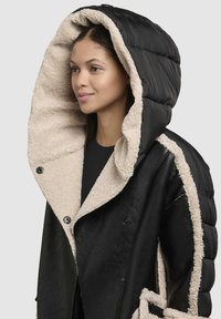 Sort og beige jakke med en fluffy indre foring, puffy hætte og dobbeltbrettede lukning med synlige knapper og en kontrasterende tekstur.